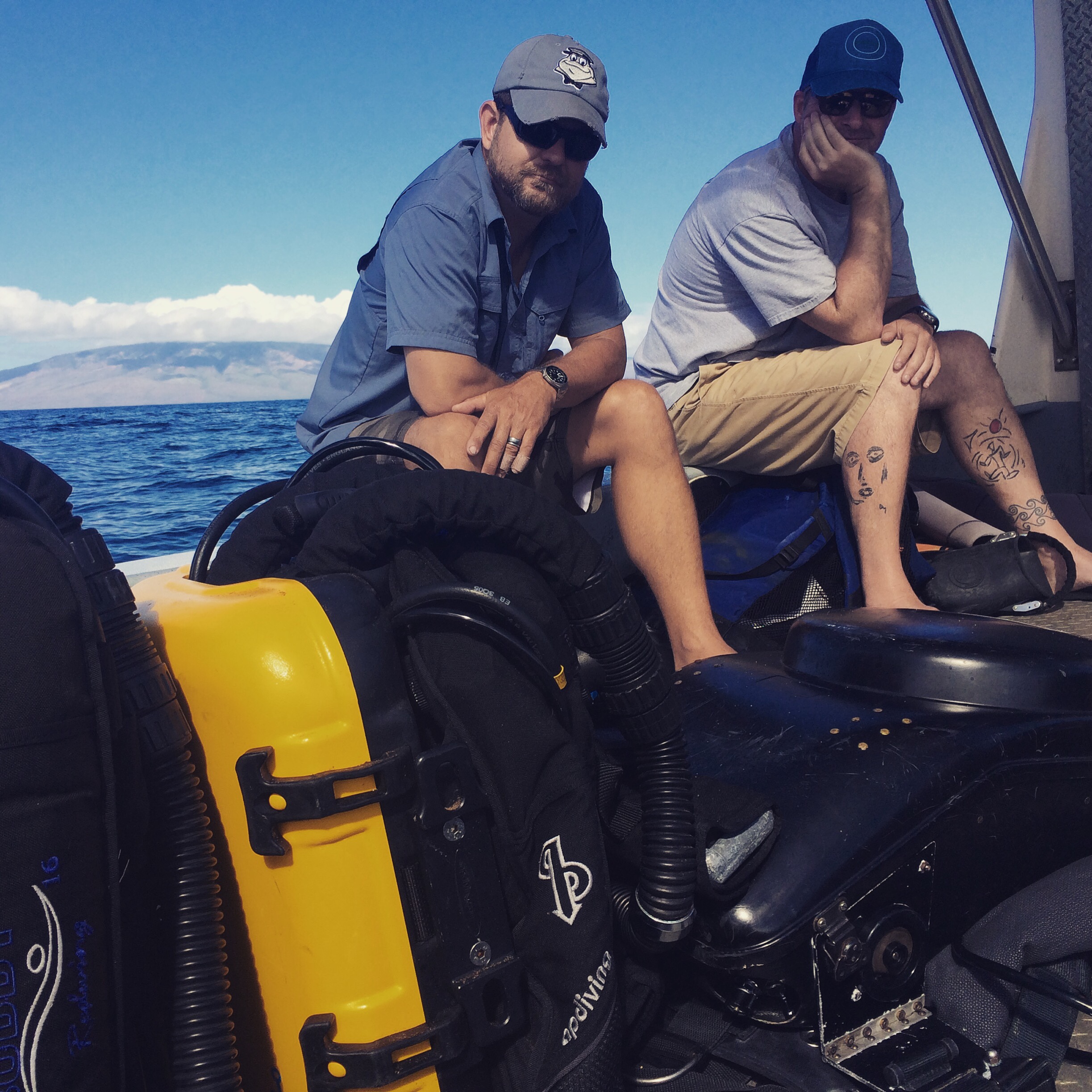 Two divers sitting on a boat with rebreather gear, Maui coastline behind them.