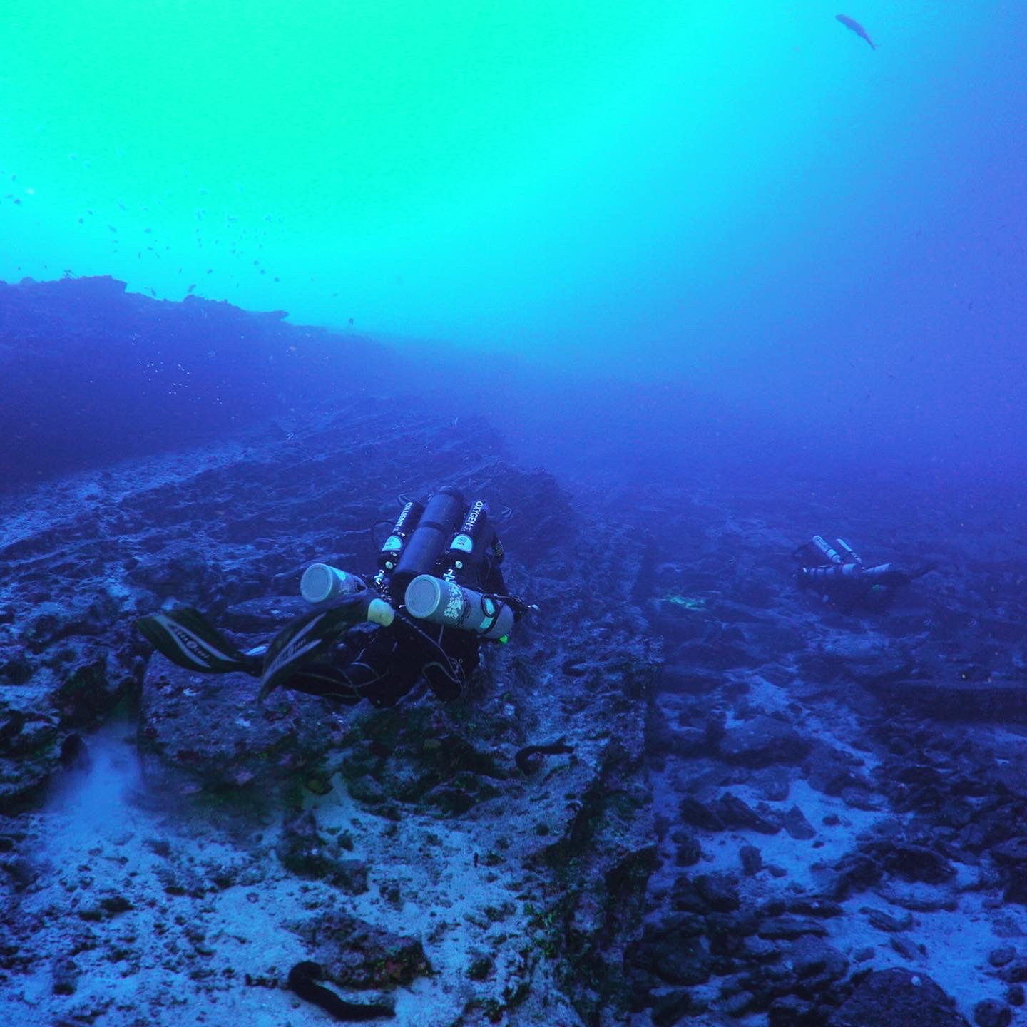 Rebreather diver over a dark seabed in deep blue water.