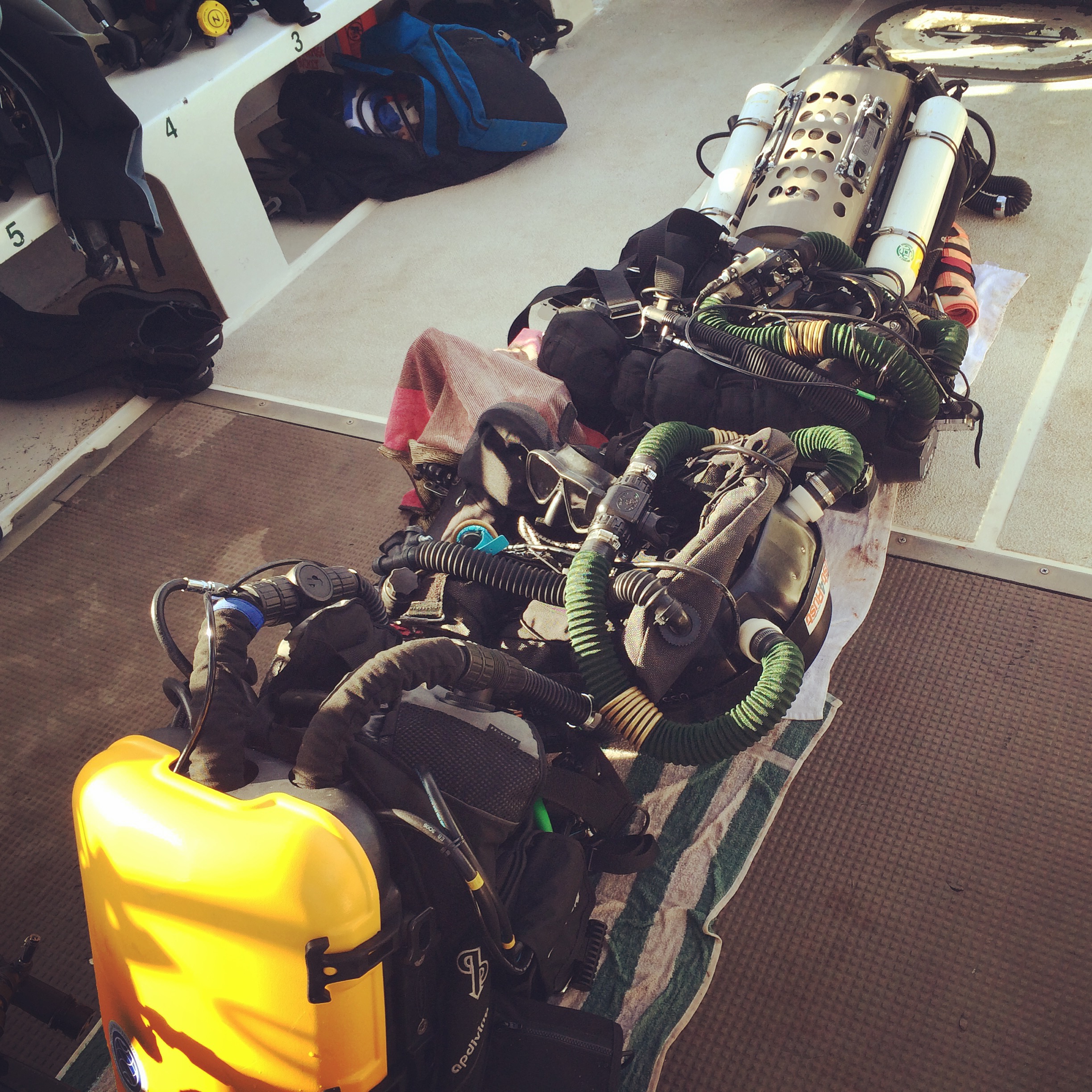 Rebreather units and dive gear lined up on a boat deck, yellow case in front.
